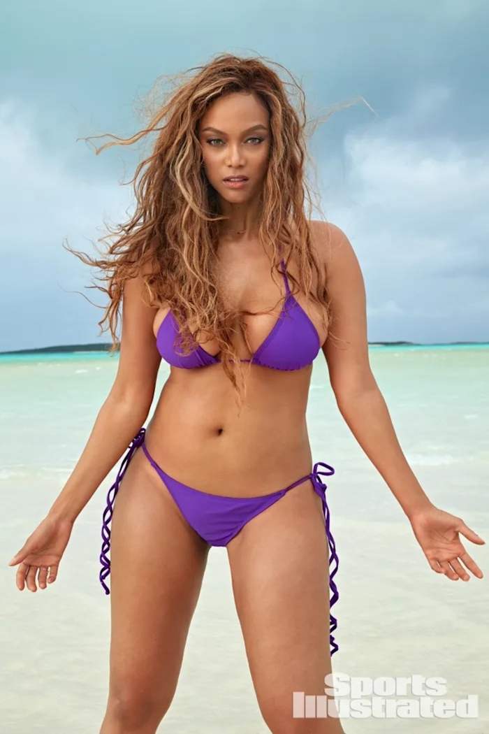 Tyra Banks stands in the shallow ocean water in a bright purple bikini set.